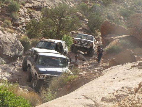 1984 Ford Bronco - Two off-road vehicles, a blue pickup truck and a white SUV, navigate a rocky, mountainous terrain. The pickup truck is in the foreground, a…