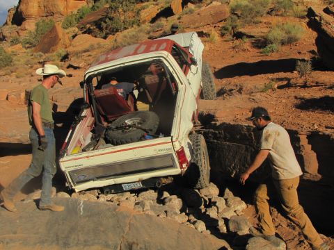 1984 Ford Bronco - Two men are working near a white and red Ford Bronco that is stuck on a rocky, off-road trail. One man in a cowboy hat is standing by the o…