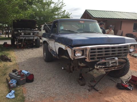 1984 Ford Bronco - Two Ford trucks are parked on a gravel lot with one in the foreground being worked on. The truck in the foreground is a blue and white pick…