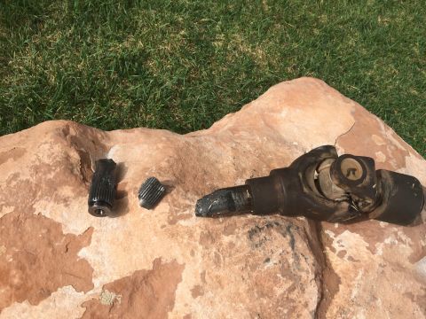 1984 Ford Bronco - Broken driveshaft components, including a universal joint and two splined shafts, are laid out on a large rock with grass in the background.