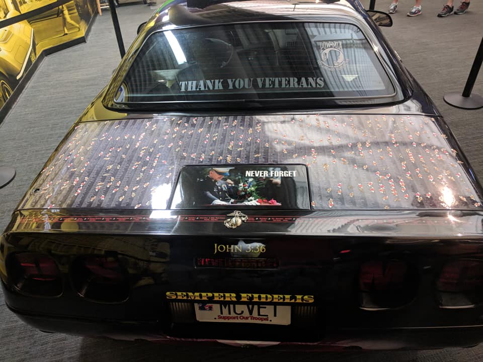 Black Chevrolet Corvette with "Thank You Veterans" on rear window and small flags covering the trunk.