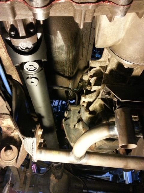 1978 Ford Bronco - Underside view of a vehicle's drivetrain, showing a black driveshaft connected to a transfer case, with exhaust piping and suspension compo…