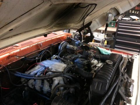 1978 Ford Bronco - The open hood of an orange 1978 Ford Bronco reveals the engine bay with the V8 engine, radiator, and various hoses and wiring.