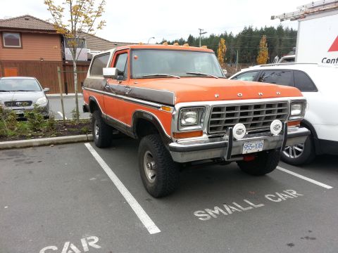 1978 Ford Bronco - A lifted, orange and brown 1978 Ford Bronco is parked in a parking spot marked "SMALL CAR". The front of the vehicle shows a chrome grille…