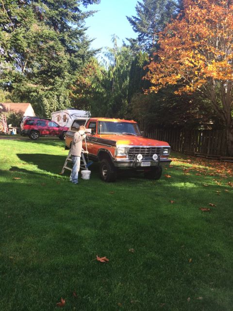 1978 Ford Bronco - A person in a grey jacket and blue jeans is washing an orange and black 1978 Ford Bronco in a grassy yard with trees in the background.