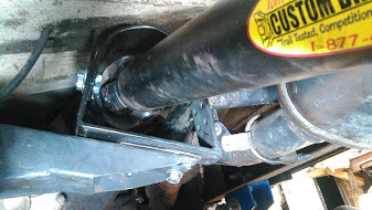 1978 Ford Bronco - A close-up, low-angle view shows a black driveshaft with a yellow and black sticker that reads "CUSTOM BUILT" and "1-877-" mounted to a bla…