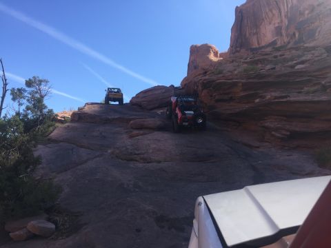 1984 Ford Bronco - Two off-road vehicles navigate a rocky, desert terrain under a clear blue sky with wispy clouds. The lead vehicle is a red Jeep-like SUV, w…