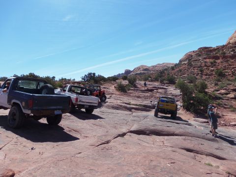 1984 Ford Bronco - Several off-road vehicles are parked on a rocky, desert trail under a clear blue sky. A modified white Ford Bronco is in the foreground, wi…