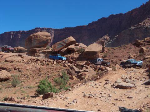 1984 Ford Bronco - Several off-road vehicles navigate a rocky, desert terrain with steep canyon walls in the background. A red SUV is visible on the left, wit…