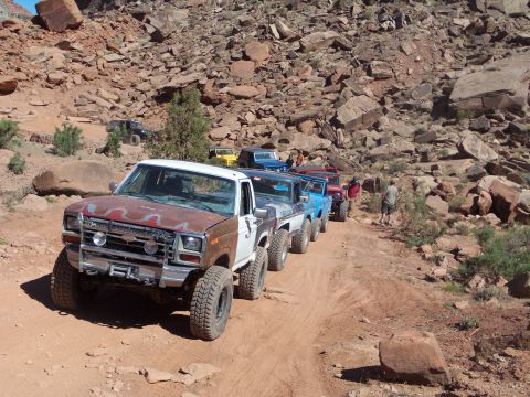 1984 Ford Bronco - A line of off-road vehicles drives down a dusty, rocky trail in a desert canyon. The lead vehicle is a white and rusty brown pickup truck w…