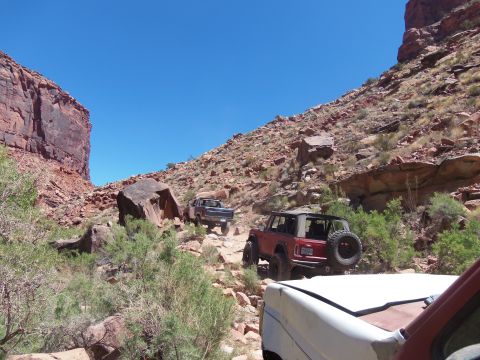 1984 Ford Bronco - Two off-road vehicles, a red Bronco and a dark-colored truck, navigate a rocky, uphill trail with steep canyon walls on either side. The Br…