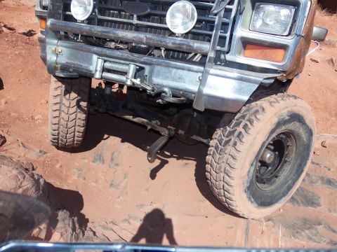 1984 Ford Bronco - The front end of a lifted 1980s Ford Bronco is shown at a steep angle, with its tires on a rocky, red dirt terrain. The vehicle features a…