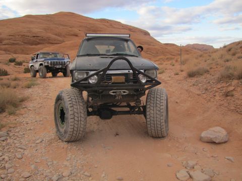 1984 Ford Bronco - Two off-road modified trucks drive on a dirt path through a desert landscape. The foreground truck is a dark-colored SUV with a prominent f…