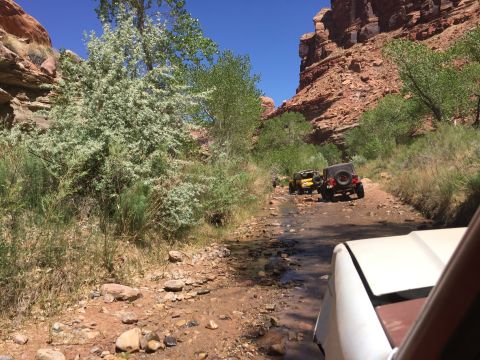 1984 Ford Bronco - A white vehicle, likely an SUV, drives down a rocky, shallow stream bed in a desert canyon. Two other off-road vehicles are visible ahead.