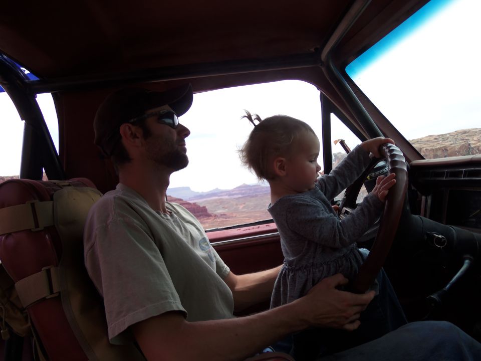 Man in sunglasses and baseball cap in driver's seat, child holds steering wheel inside a vehicle.