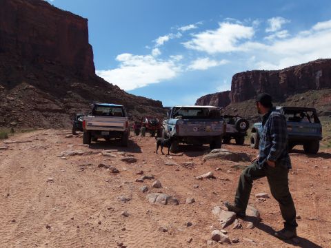 1984 Ford Bronco - Several off-road vehicles are parked on a dusty, rocky trail with large red rock formations in the background under a blue sky with scatter…