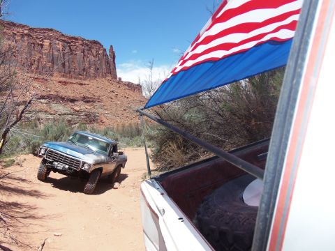 1984 Ford Bronco - A blue and silver 1970s Ford F-Series pickup truck is driving off-road on a dirt path with a large red rock formation in the background. An…