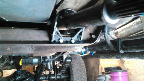 1978 Ford Bronco - The underside of a vehicle shows a driveshaft supported by a custom metal bracket attached to the frame. Visible components include leaf sp…