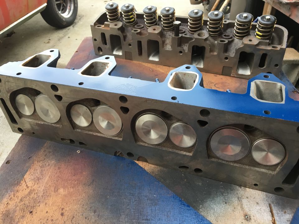 Two Ford V8 engine cylinder heads, one blue, on a wooden surface.