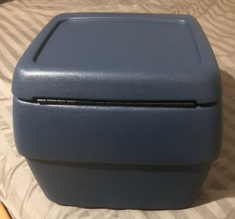 1995 Ford Bronco - A blue plastic center console storage box with a hinged lid is shown from a front-facing angle. The box rests on a striped fabric surface.