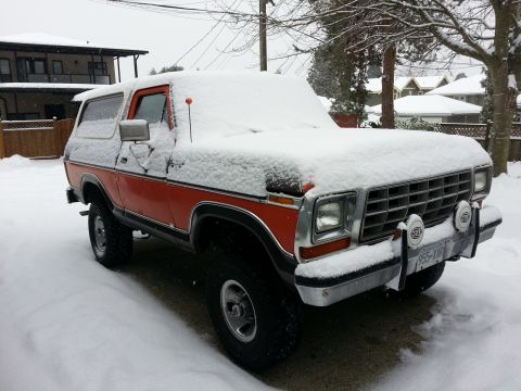 1978 Ford Bronco - A classic orange and black Ford Bronco is parked outside in the snow, partially covered in snow. The vehicle is viewed from the front three…