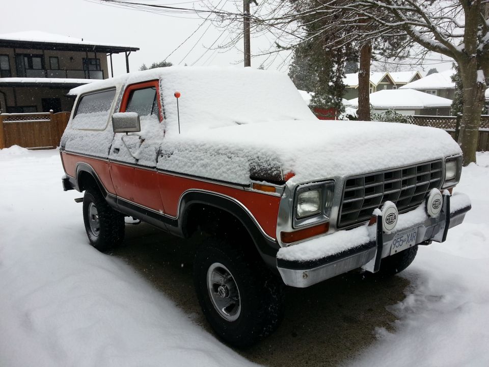 Orange and black Ford Bronco covered in snow, parked in a snowy driveway.