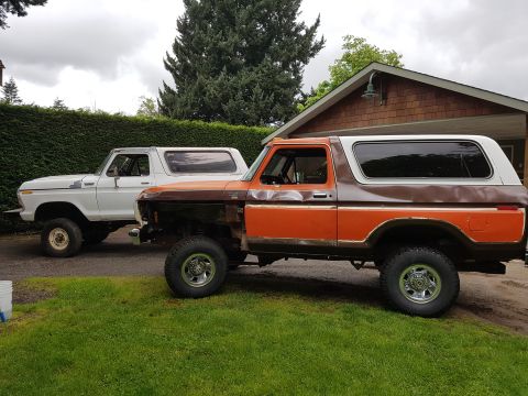 1978 Ford Bronco - Two Ford Broncos are parked side-by-side in a grassy yard next to a house. The Bronco on the left is white and the Bronco on the right is o…