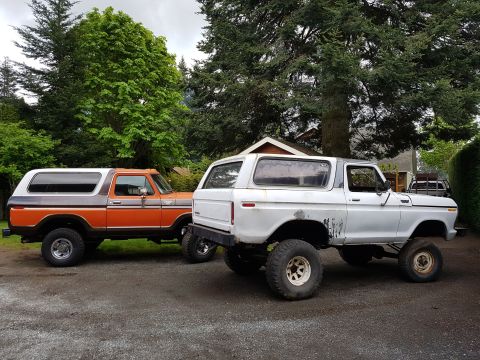 1978 Ford Bronco - Two lifted Ford Broncos are parked side-by-side on a gravel surface in front of trees and a house. The Bronco on the left is orange and bro…
