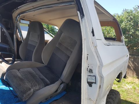 1978 Ford Bronco - The interior of a white Ford Bronco is shown, featuring grey striped bucket seats and a roll cage. The driver's side door is open, revealin…