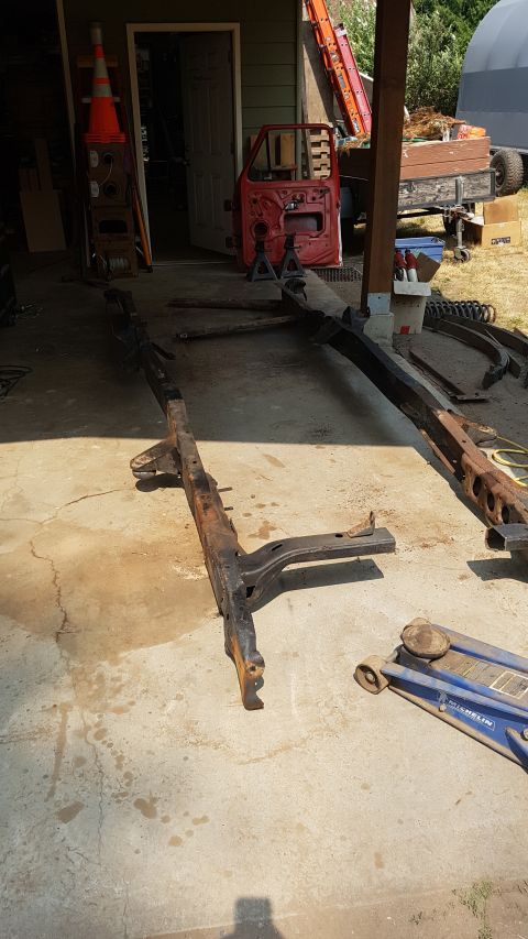 1978 Ford Bronco - Two rusty vehicle frame rails lie on a concrete surface in front of a garage. A red vehicle door is propped up on jack stands to the left o…