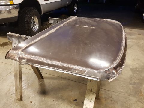 1978 Ford Bronco - stripped bare-metal truck hood with a center crease sits on sawhorses in a garage, with a van and alloy wheel in the background.