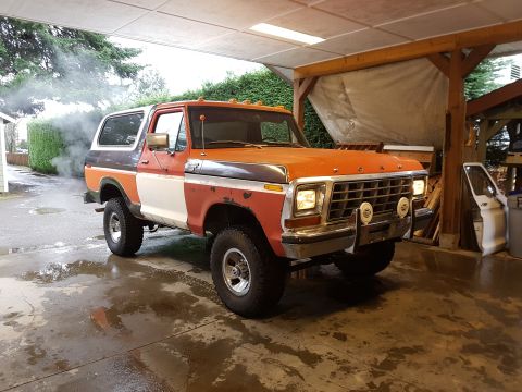 1978 Ford Bronco - A vintage Ford Bronco with a tri-color paint scheme (orange, white, and dark brown/grey) is parked under a covered carport. Smoke is billow…