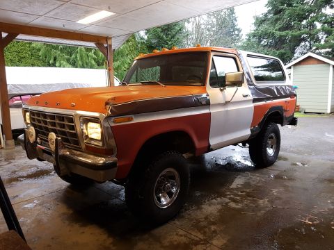 1978 Ford Bronco - A three-quarter front view of a vintage Ford Bronco with an orange, brown, and white paint scheme. The vehicle is parked under a covered ca…