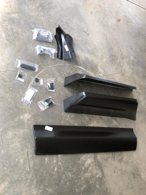 1978 Ford Bronco - A collection of black metal body panels and small hardware parts are laid out on a concrete surface. The parts appear to be for a vehicle,…