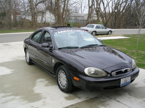1996 Mercury Sable - A dark sedan with a "MERCURY" banner on the windshield is parked on a wet driveway, with another car in the background.