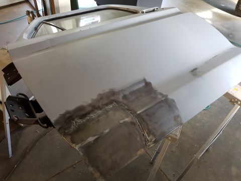 1978 Ford Bronco - A gray vehicle door is shown supported by sawhorses, with visible metal repair work and welding on the lower section. The door appears to b…