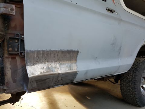 1978 Ford Bronco - The lower portion of a light gray vehicle door is shown, featuring freshly welded metal repair work along the bottom edge. The vehicle appe…