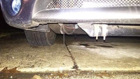 Underside of a car's front bumper with a tire visible and liquid dripping onto concrete.