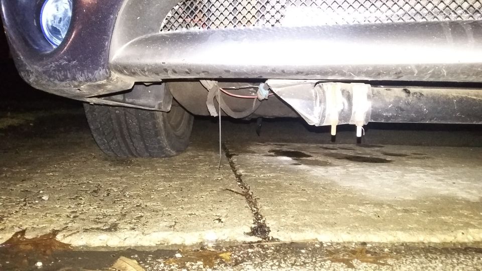 Underside of a car's front bumper with a tire visible and liquid dripping onto concrete.