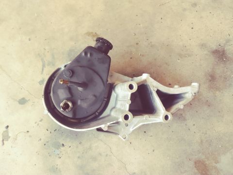 1996 Ford Bronco - A power steering pump assembly with a metal bracket is shown from a top-down perspective, resting on a concrete surface.