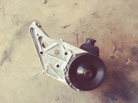 1996 Ford Bronco - A power steering pump assembly with a pulley and reservoir is mounted on a silver bracket. The assembly is placed on a concrete surface wit…
