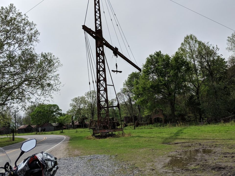 Motorcycle parked on gravel road next to a large rusted crane and old buildings.