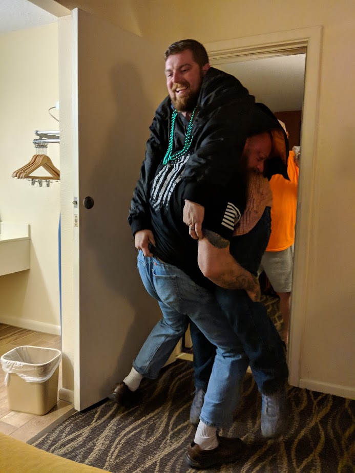 Two men in a hotel room doorway, one carrying the other on his shoulders.