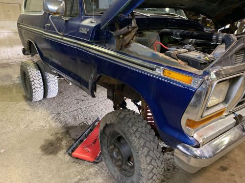 1979 Ford Bronco - The driver's side of a dark blue 1979 Ford Bronco is shown with the hood open, revealing the engine bay. The vehicle has dual rear wheels a…