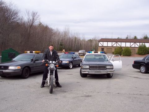 2001 Ford Crown Victoria - A man sits on a motorcycle in front of several police cars, one with its door open.