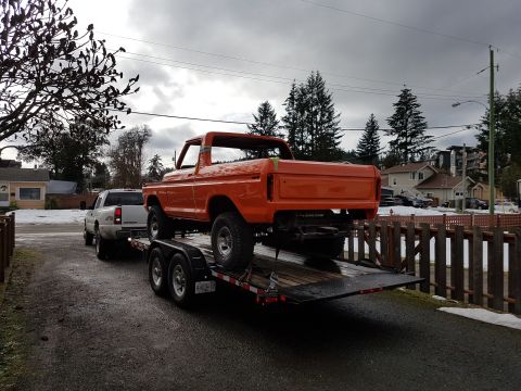 1978 Ford Bronco - Orange 1978 Ford Bronco on a trailer, towed by a white pickup truck on a cloudy day.