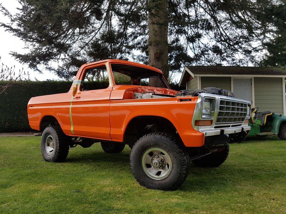 Orange pickup truck with hood open, parked on grass. A green riding mower is visible in the background