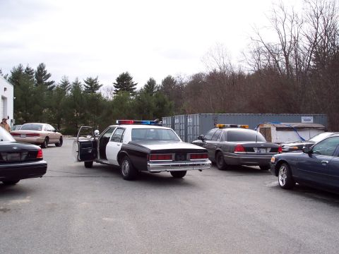 2001 Ford Crown Victoria - Several police vehicles and civilian cars are parked in an outdoor lot under an overcast sky.