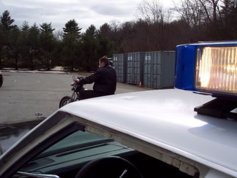 2001 Ford Crown Victoria - A close-up view of a police car's light bar, with a man riding a motorcycle in the background.