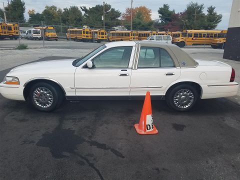 2003 Mercury Grand Marquis - A white 2003 Mercury Grand Marquis sedan with a tan roof is parked on asphalt, with school buses in the background.